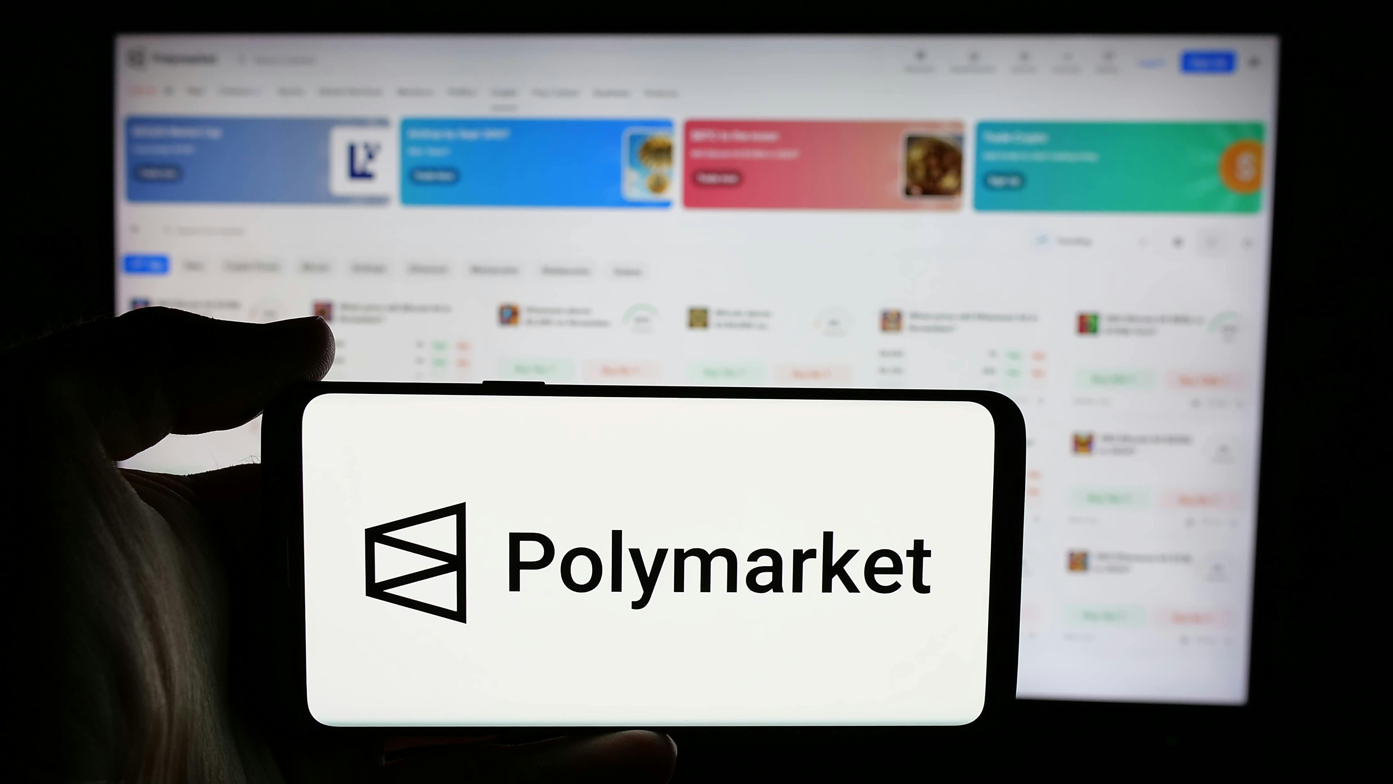 In this photo illustration, a person is holding a cellphone with the logo of crypto prediction market company Polymarket on screen in front of business webpage.
